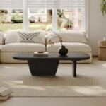 A modern black wood coffee table with an asymmetrical base sits on a light textured rug in front of a cream sofa near large windows.