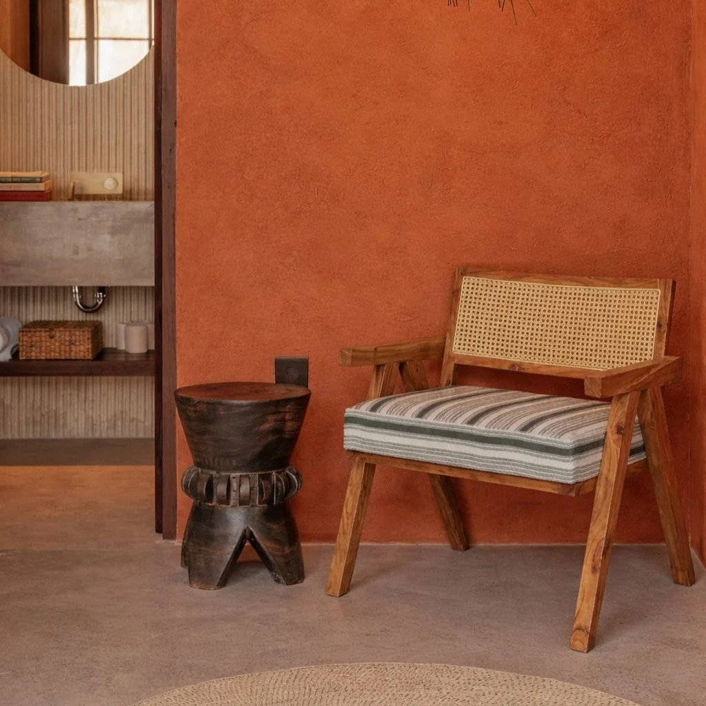 Cane wooden lounge chair with striped cushion placed against a terracotta wall with hanging lamp and rustic decor.