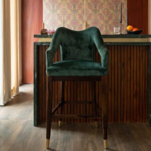 Emerald green velvet-style upholstered bar chair with curved backrest, cushioned armrests, dark wooden legs, footrest, and gold-finished leg caps.