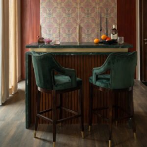 Emerald green velvet-style upholstered bar chair with curved backrest, cushioned armrests, dark wooden legs, footrest, and gold-finished leg caps.