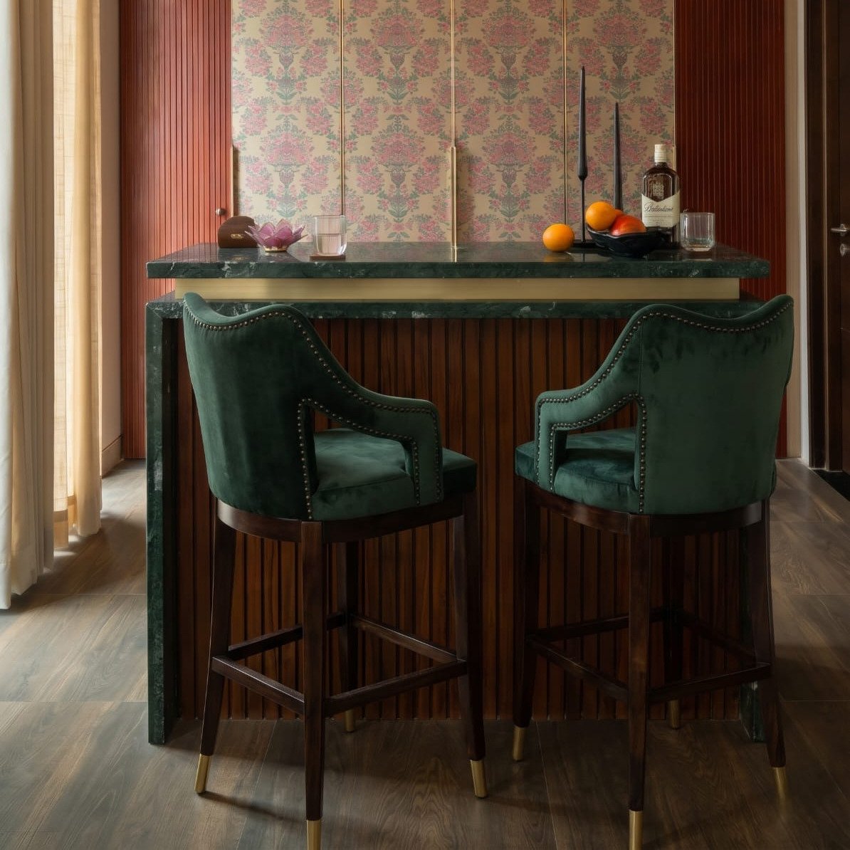 Emerald green velvet-style upholstered bar chair with curved backrest, cushioned armrests, dark wooden legs, footrest, and gold-finished leg caps.